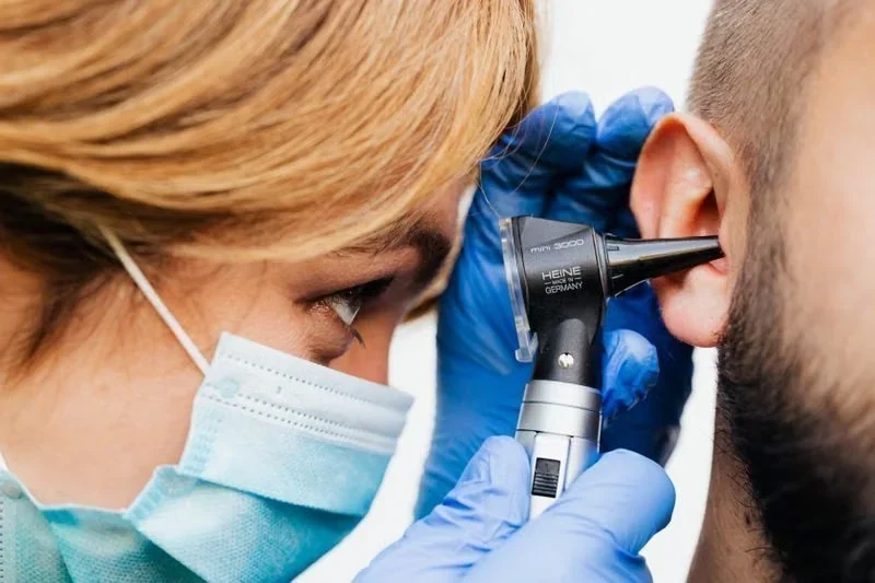 Profissional de saúde com máscara e luvas examinando o ouvido de um paciente com otoscópio