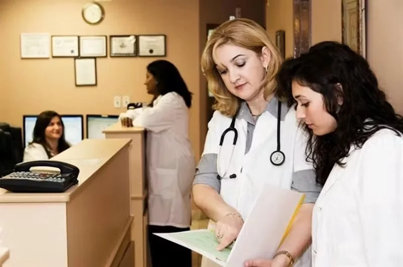 Duas médicas em jalecos brancos analisam prontuário, com recepcionistas em balcão de clínica ao fundo