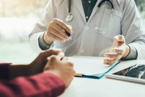Médico de jaleco e estetoscópio explica a paciente, apontando documento com caneta. Tablet na mesa
