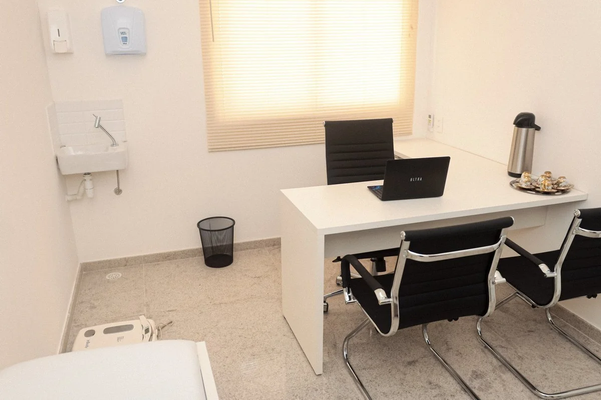 Consultório médico com mesa, cadeiras, laptop, pia, dispensadores na parede, balança e janela com persiana