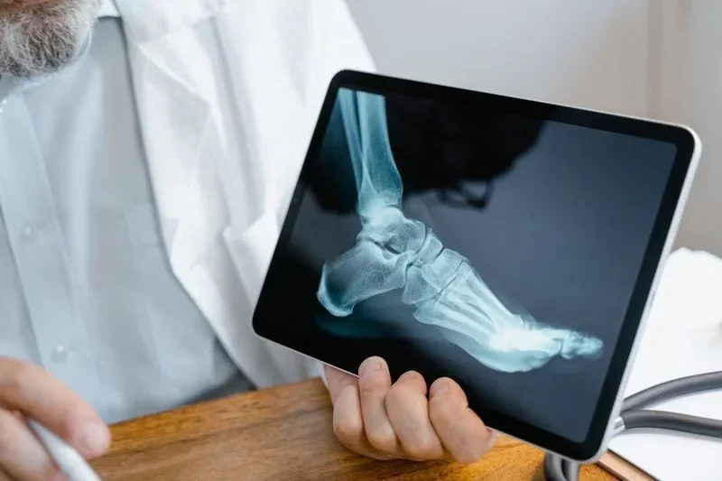 Médico com barba grisalha segurando tablet com raio-x de pé, usando jaleco branco e caneta em mesa de madeira