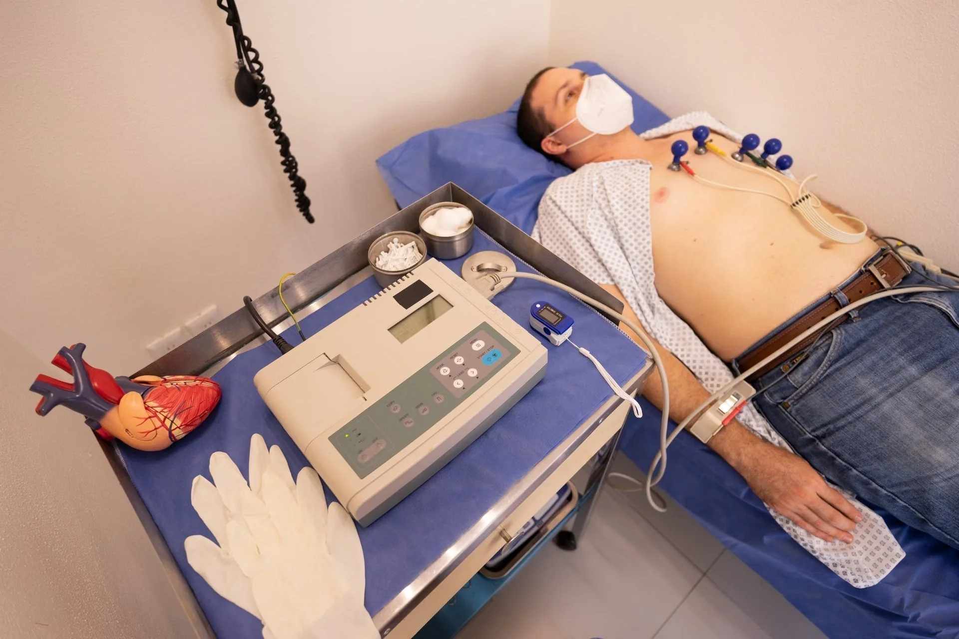 Homem deitado em maca com eletrodos de ECG no peito e oxímetro no dedo. Máquina de ECG, luvas e modelo de coração