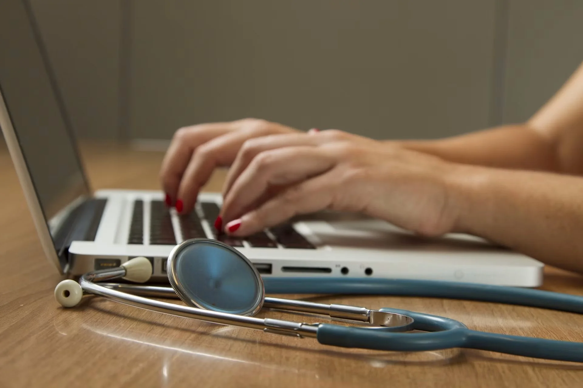 Mãos digitando em laptop, com estetoscópio azul em primeiro plano sobre mesa de madeira