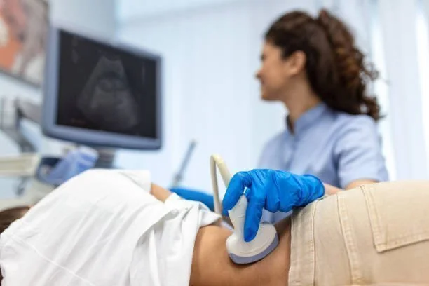 Profissional de saúde com luva azul realizando ultrassom no abdômen de paciente, monitor de ultrassom ao fundo