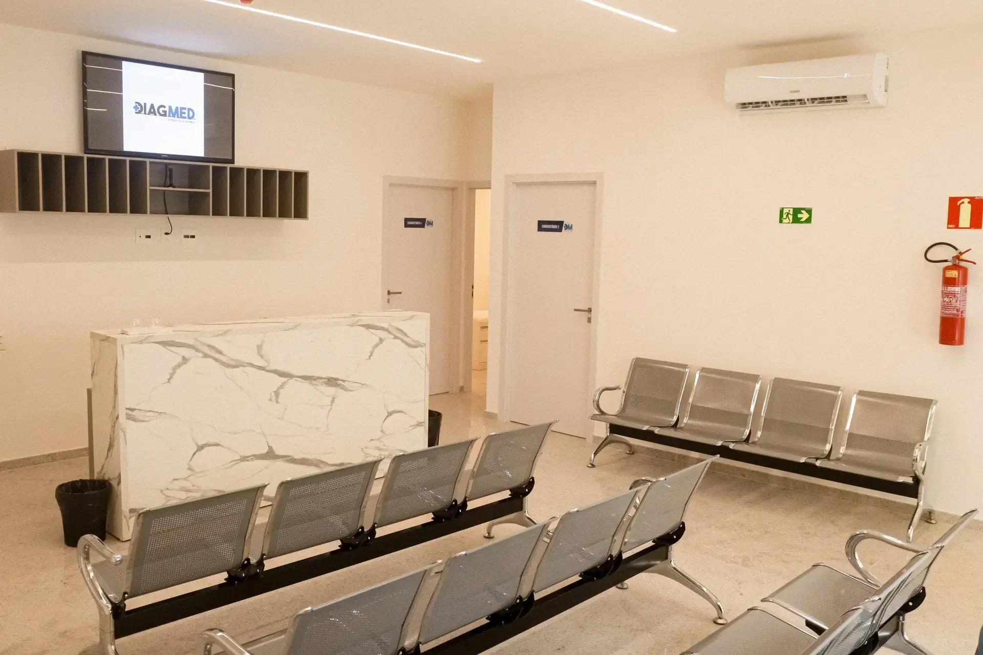 Sala de espera de clínica com balcão de recepção, TV com logo Diagmed, cadeiras metálicas, ar condicionado e extintor
